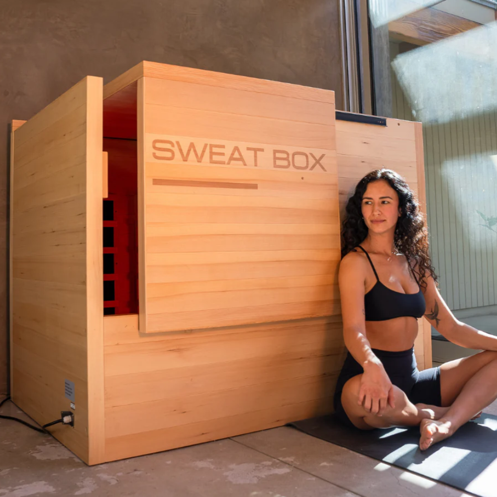 A woman relaxing next to the Sweat Box Infrared Sauna, designed for deep detoxification, red light therapy, and home wellness benefits.