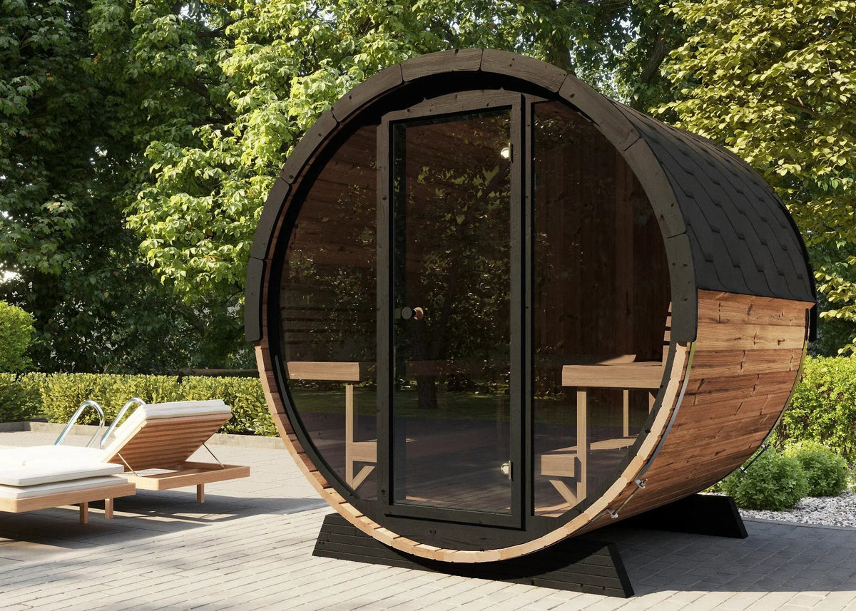 The SaunaLife EE8G Barrel sauna with a dark wood exterior, black metal frame, and large glass doors, shown on a paved patio with lounge chairs and a lush green garden background.