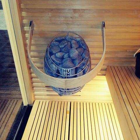 HUUM DROP 9kW Electric Sauna Heater with a rounded stainless steel basket filled with dark stones, mounted on a wooden sauna wall