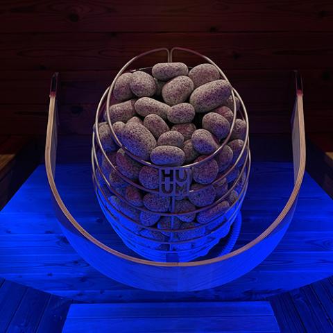HUUM DROP electric sauna heater studio shot with rounded stone basket and blue accent lighting, front facing view